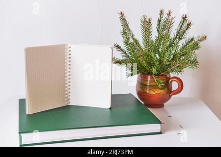 Blank sheets of opened checkered notepad on green book, bouquet of christmas tree branches in handmade clay mug. New Year's planning, goal setting, mo Stock Photo