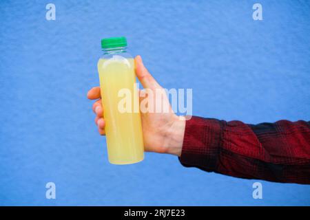 Close-up hand of unrecognizable man in red shirt holding Lemonade Plastic Bottle in front of Blue Wall Background in Outdoors Stock Photo