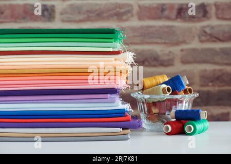 Stack of colorful monochromatic fabrics and spools of thread on brick wall background Stock Photo