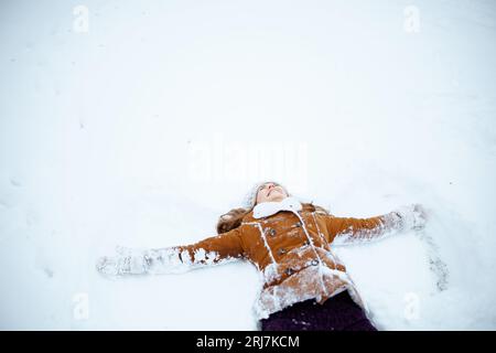 smiling modern middle aged woman in brown hat and scarf in sheepskin coat with mittens making snow angels outside in the city in winter. Stock Photo