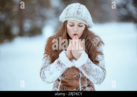 elegant middle aged woman in brown hat and scarf in sheepskin coat warming cold hands with breath outside in the city in winter. Stock Photo