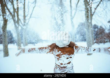 Seen from behind woman in brown hat and scarf in snowy clothes and sheepskin coat with mittens catching snow outside in the city in winter. Stock Photo