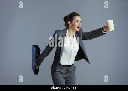 smiling modern female employee in gray suit with coffee cup and briefcase running isolated on gray background. Stock Photo