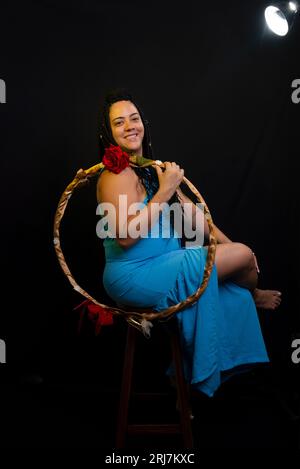 Beautiful woman wearing blue colored outfit sitting holding a hula hoop against black background in studio. Positive person. Stock Photo