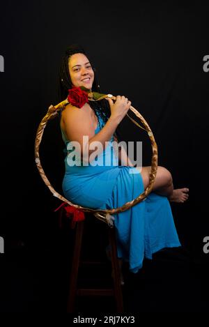 Beautiful woman wearing blue colored outfit sitting holding a hula hoop against black background in studio. Positive person. Stock Photo