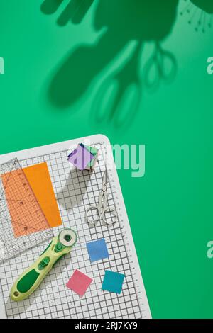 Rotary cutter, ruler, bright square pieces of fabric, stack of bright fabric square pieces, scissors on craft mat, green background Stock Photo