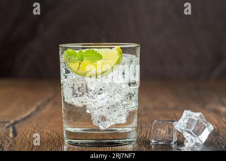 Mineral refreshing water in a glass with ice, mint leaves and lime slice. Dark wooden surface. Artificial ice cubes. Stock Photo