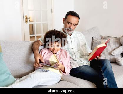 Hispanic Grandson Using Tablet Computer While Grandpa Reading Book Indoor Stock Photo