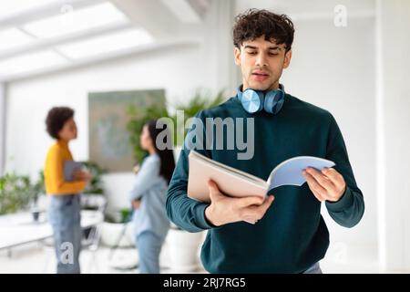 Serious college student guy holding open copybook in hands, standing in modern audience with classmates on background Stock Photo