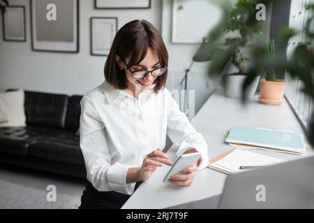 Young female employee using cellphone at workplace in office or coworking space, enjoying break at work, free space Stock Photo