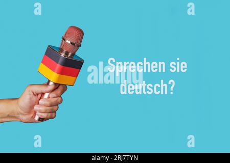 the question do you speak german written in german and the hand of a man holding a microphone patterned with the german flag against a blue background Stock Photo