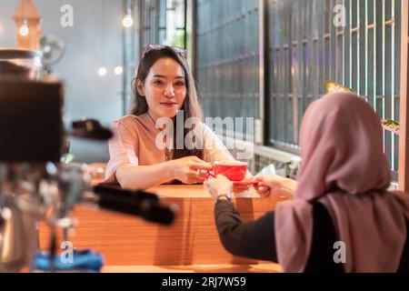 A beautiful Asian barista handing a cup of coffee to customers who wait for orders at the coffee shop Stock Photo