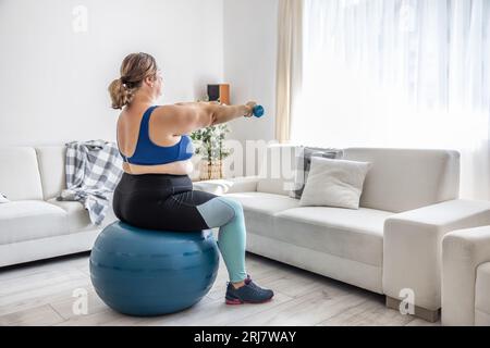 Overweight lady doing sports exercise in living room. He sits on a fitness ball and holds his hands with dumbbells in front of him. Stock Photo