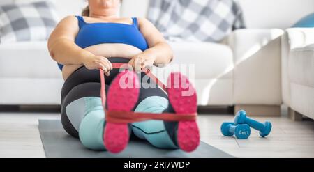 Overweight lady doing sports exercise in living room. He lies on the mat and stretches the sports rubbers. Stock Photo