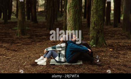 Tired cheerful young caucasian couple in jackets sits in forest on mat, enjoy travel at weekend, rest, relax Stock Photo
