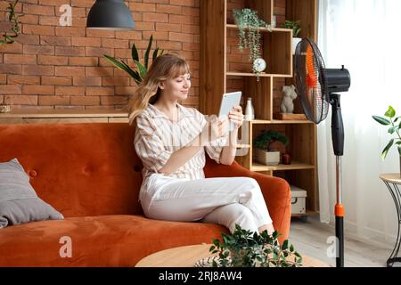 Young woman with tablet computer and blowing electric fan sitting on sofa at home Stock Photo