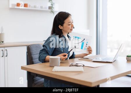 Side view of attractive woman indicating on graphs while holding video conference via laptop and headset at home. Pretty business lady analyzing sales Stock Photo