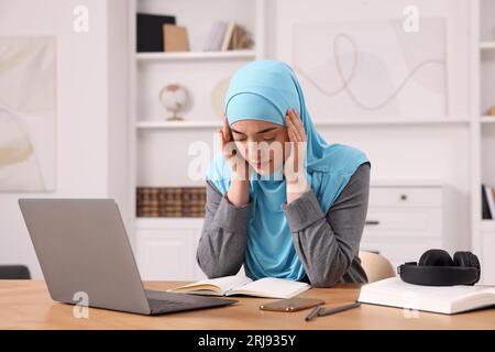 Tired Muslim woman working near laptop at wooden table in room Stock Photo