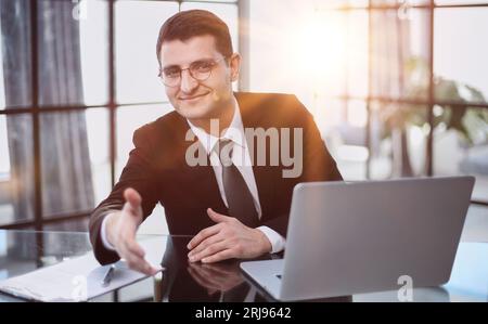 working at the computer, makes an inviting gesture, offers to sit down for an interview Stock Photo