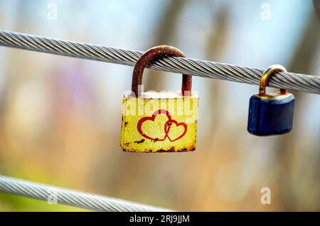 Old rusty yellow padlock with two hearts hanging on a metal fence next to small usual padlock outdoors Stock Photo