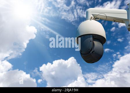 Closeup of a modern white and black security camera against a clear blue sky with clouds, sunbeams and copy space. Stock Photo