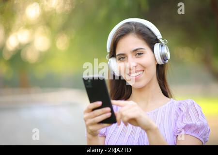 Happy female wearing headphone and holding phone listening to music looking at camera in a park Stock Photo