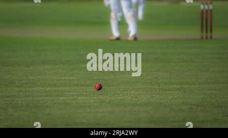A red cricket ball, inches above the grass and heading for the boundary.  The stumps and part of a player are visible in the out of focus background Stock Photo