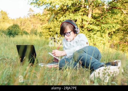 Concentrated junior pupil in glasses and earphones lying on green grass in park in front of laptop and doing homework in notebook. Boy with dark hair Stock Photo