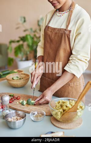 Vertical closeup of young black woman cutting fresh vegetables while cooking pasta dish in cozy kitchen Stock Photo