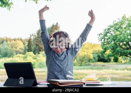 Boy with earphones, glasses sitting at table where standing tablet and lying notebooks, stretching arms after studying. Hard-working pupil in gray lon Stock Photo