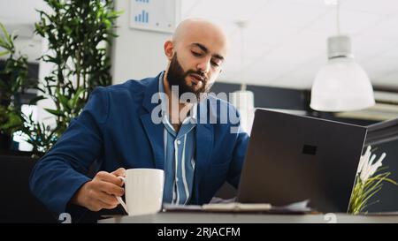 Middle eastern man reads documents online before creating professional marketing report in office. Male worker trying to find business solution, entrepreneur in coworking space. Stock Photo