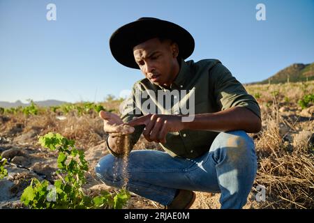Young black man in brimmed hat crouching and examining dirt next to plants on farm Stock Photo