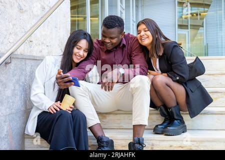 Happy coworkers taking a selfie photo at work break. Group of young student friends smiling at university outdoors. Ethnic diversity. Stock Photo