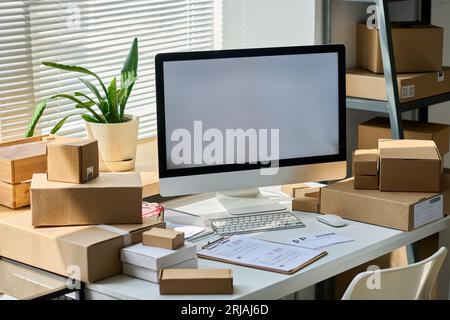 Desktop computer with blank screen standing among packed boxes containing orders of online clients on workplace of warehouse manager Stock Photo