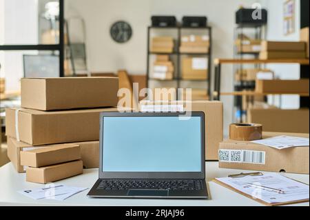 Laptop with blank screen standing among documents and packed boxes containing orders of online shops clients in storage room Stock Photo