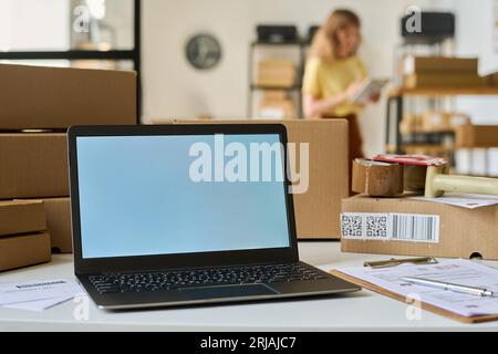 White screen of laptop standing on desk among packed cardboard boxes and financial documents on background of young female manager Stock Photo