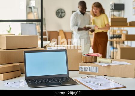 Laptop with white empty screen standing on desk among packed boxes and documents against two young intercultural managers using tablet Stock Photo