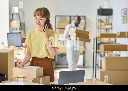 Young smiling female manager of warehouse speaking to client on mobile phone and writing down personal information on top of packed box Stock Photo