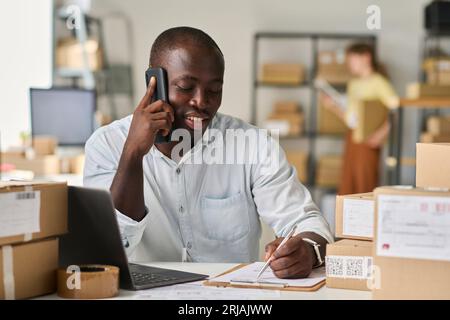 Happy young male manager of marketplace talking to client on mobile phone and making notes in document while sitting in front of camera Stock Photo