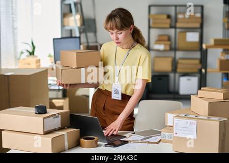 Young woman in casualwear holding stack of packed boxes and using laptop while checking personal data of customer in database Stock Photo
