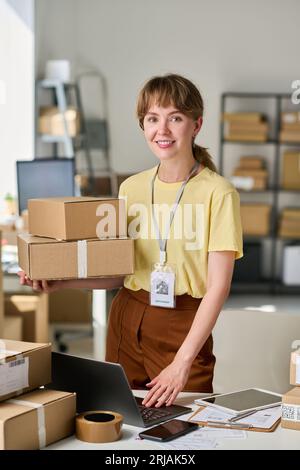 Happy young female owner of warehouse lookng at camera by workplace with laptop while holding stack of boxes and checking online data Stock Photo