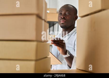 Young serious African American manager of warehouse with document looking at stack of packed boxes while standing in front of shelf Stock Photo