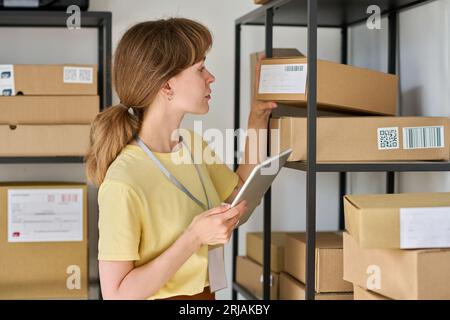 Side view of young female owner of marketplace checking personal data about clients on packed boxes while standing by rack with orders Stock Photo