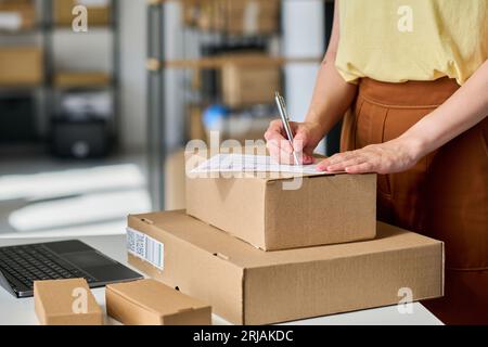 Close-up of young female worker of warehouse writing down personal data on consignment on top of stack of boxes in front of camera Stock Photo
