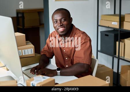 Happy young African American man in brown shirt looking at camera while sitting by workplace in warehouse and typing on computer keyboard Stock Photo