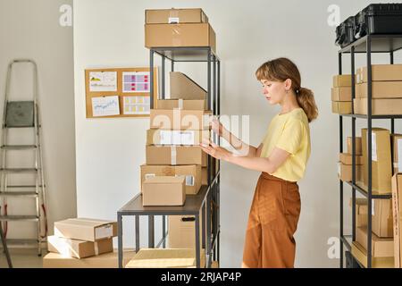 Side view of young pretty manager of storage room standing in front of rack with parcels and looking through numbers of orders on boxes Stock Photo