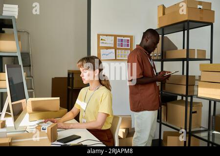 Side view of two young intercultural workers of warehouse checking data while man using tablet and woman networking by computer Stock Photo