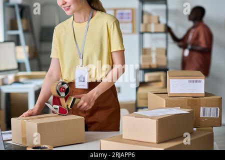 Cropped shot of young female manager of warehouse preparing parcels for sending to customers while sealing boxes with cellotape Stock Photo