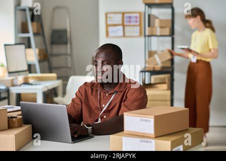 Young serious African American man typing on laptop against female colleague while checking information about customers and goods Stock Photo