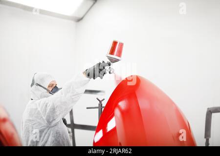 Male worker in protective clothes and mask painting car using spray paint. Stock Photo